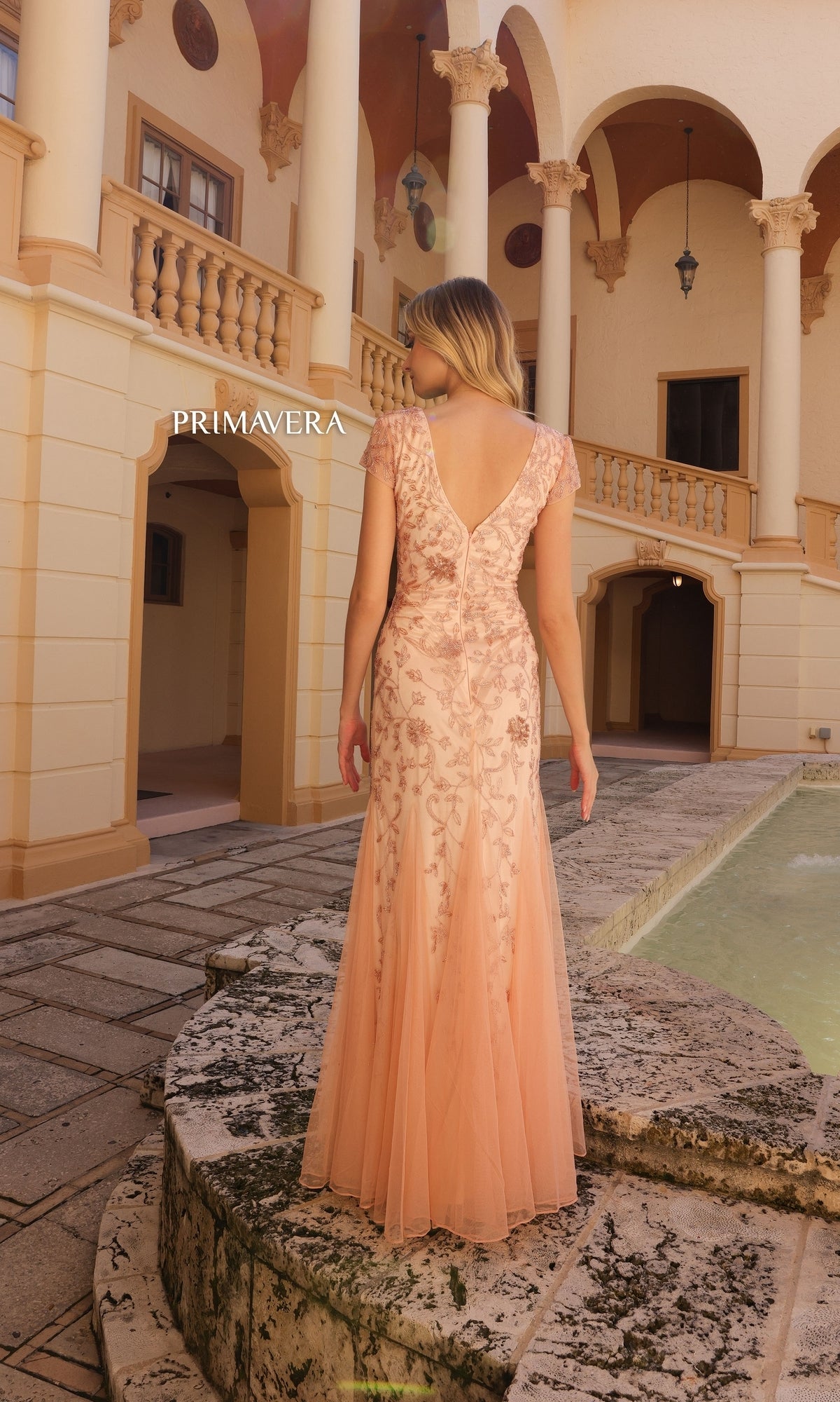 A woman stands with her back to the camera in a courtyard, wearing the Long Prom Dress 13112 by Primavera—a peach floral beaded evening gown with a v-neckline.